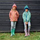 Two children wearing hats and patterned raincoats, standing against a black wooden wall in green boots and trousers in front of grass.